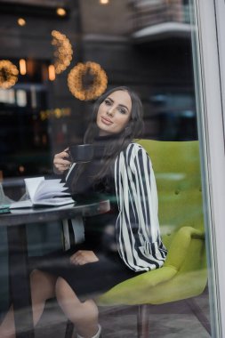 A beautiful young woman in a black dress sits in a cafe by the window and drinks coffee. Coffee break.