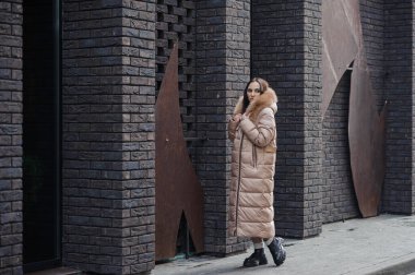 Beautiful young pregnant woman in a brown coat, in the background a brick wall.