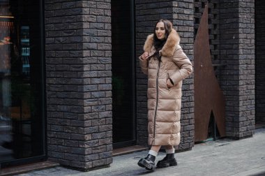 Beautiful young pregnant woman in a brown coat, in the background a brick wall.
