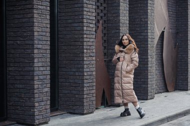 Beautiful young pregnant woman in a brown coat, in the background a brick wall.