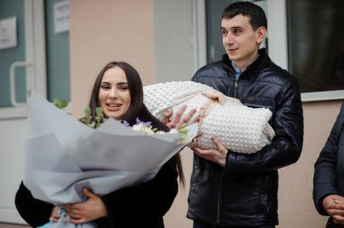 Bir erkek, elinde çiçek tutan güzel bir kadının yanında battaniyeye sarılı bir bebek tutar.