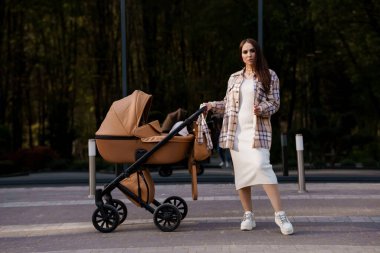 Güzel bir kızın portresi. Ekose gömlekli ve şapkalı güzel bir kızın yakın plan fotoğrafı. Kız kahverengi bebek arabasının yanında duruyor. Anne küçük bir çocukla yürüyor.