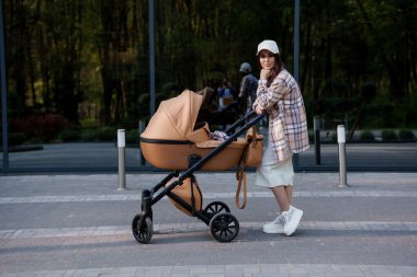 Güzel bir kızın portresi. Ekose gömlekli ve şapkalı güzel bir kızın yakın plan fotoğrafı. Kız kahverengi bebek arabasının yanında duruyor. Anne küçük bir çocukla yürüyor.