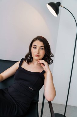 Siyah elbiseli güzel bir kızın portresi beyaz arka planda bir sandalyede oturuyor.