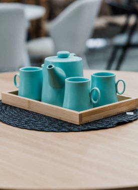 Sarı ahşap masa, bej ahşap koltuklar ve yumuşak kumaş döşeme. Masada mavi porselen bir çaydanlık ve mavi kupalar var.