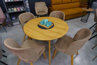 Sarı ahşap masa, bej ahşap koltuklar ve yumuşak kumaş döşeme. Masada mavi porselen bir çaydanlık ve mavi kupalar var.