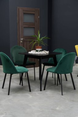 Siyah ahşap masa ve yumuşak kumaş döşemeli yeşil ahşap sandalyeler. Masanın üzerinde kahverengi bir saksıda yeşil bir bitki var.
