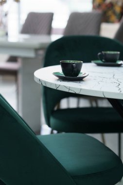 Beyaz ahşap masa, yumuşak kumaş döşemeli yeşil ahşap sandalyelerle tamamlanmış. Yeşil fincanlar ve tabaklar masanın üstünde.