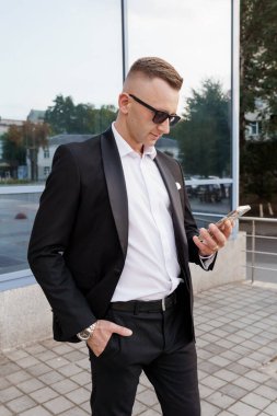 Siyah takım elbiseli şık bir adam şehir merkezinde telefonunu kontrol ediyor..