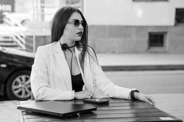 Stylish Businesswoman in Striped Blazer and Sunglasses Sitting at Outdoor Cafe Table with Laptop. clipart