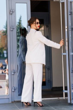 Elegant Business Woman Exiting a Modern Building in Striped Suit with Sunglasses. clipart