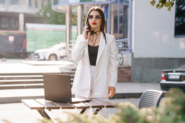 Stylish Businesswoman in Striped Suit Talking on Phone with Laptop Outdoors.