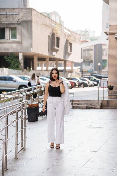 Стильная городская женщина в полосатых штанах и черном топе уверенно прогуливается по современному городу..