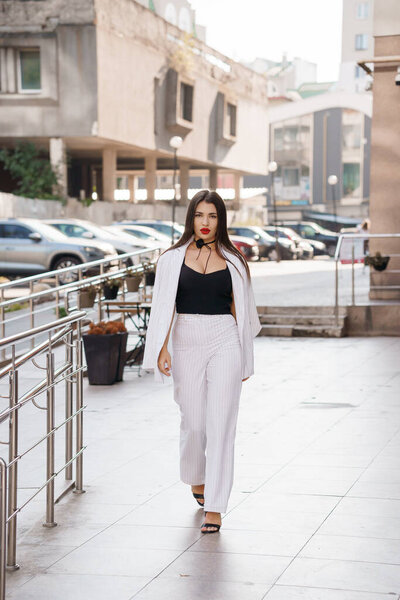 Элегантная женщина в шикарном деловом наряде уверенно прогуливается в городской обстановке.