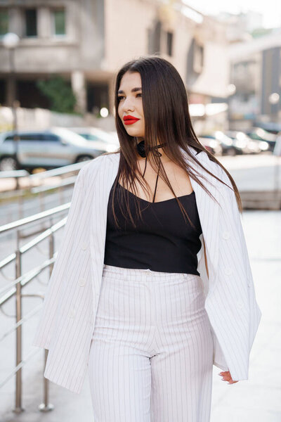 Стильная молодая женщина с длинными волосами в модном костюме в полоску и красной помадой уверенно гуляет в городской обстановке.