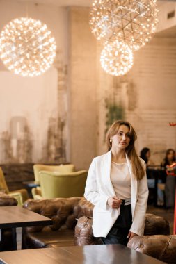 Şık ceketli zarif bir kadın modern bir kafede sanatsal ışıklandırmayla duruyor..