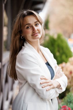 Profesyonel bir kadın, kendine güvenen bir gülümsemeyle şık beyaz bir ceketle dışarıda poz veriyor..