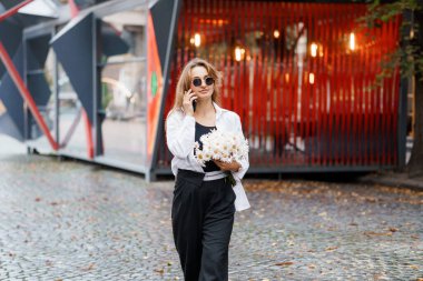 Stylish Young Woman in Sunglasses Holding Flowers While Walking in a Modern Urban Setting. clipart