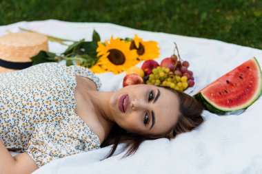 Etrafı meyveler ve ayçiçekleriyle çevrili bir battaniyede dinlenen genç kadın..