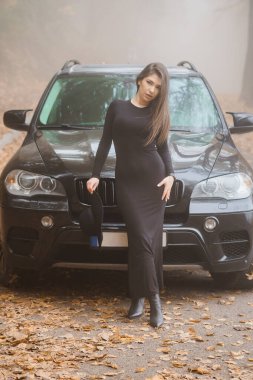 Siyah Elbiseli Zarif Kadın, Sisli Sonbahar Peyzajı Ortasında Lüks SUV 'un Önünde Emin Bir Poz Veriyor.