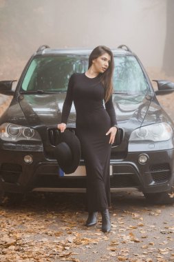 Enigmatic Beauty in the Mist: A Fashionable Woman Poses by a Black SUV in Autumn Leaves. clipart