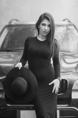 Elegant Woman in Black Dress Posing with Hat in Front of SUV. clipart