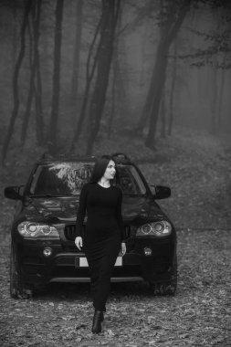 Mysterious Woman in Black Dress Standing Before SUV in Foggy Forest. clipart