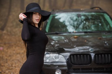 Gizemli Zarafet: Siyah Şapkalı Bir Kadın Sisli Ormanda Bir Cipin Yanında Poz Veriyor.