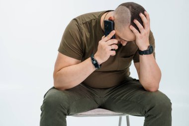 Man in olive green shirt sitting on a chair, stressed while speaking on a phone. clipart