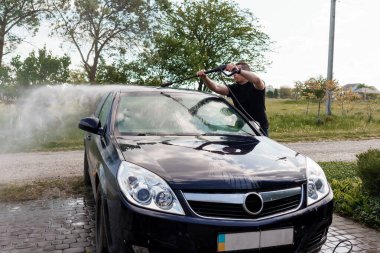 Siyah bir arabayı güneş ışığında su hortumuyla yıkayan adam..
