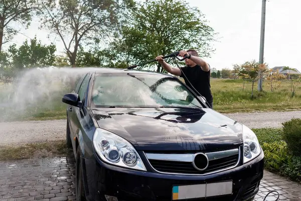 Siyah bir arabayı güneş ışığında su hortumuyla yıkayan adam..