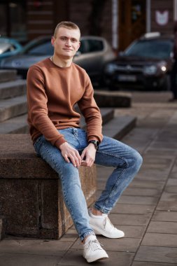 Sıradan giyinmiş genç bir adam beton merdivenlerde, şehir zemininde oturuyor..