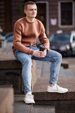 Genç adam sıradan kıyafetlerle merdivenlerde oturuyor. Elinde kahve fincanı var..