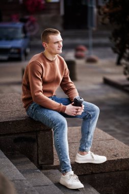 Genç adam kahve fincanı ile taş basamaklarda oturuyor, kahverengi kazak ve kot pantolon giyiyor..