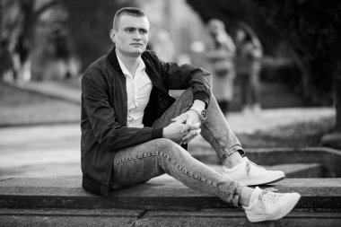 Parkta oturan, doğal ve yumuşak ışıkla çevrili, günlük kıyafetli genç bir adam..