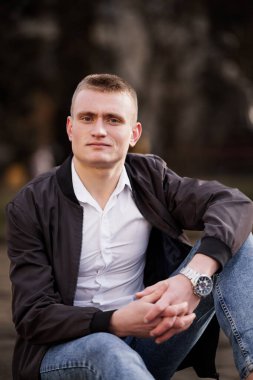 Açık havada oturan genç adam, yansıtıcı bir ifadeyle günlük kıyafetiyle..