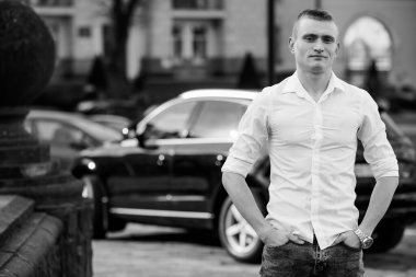 Beyaz tişörtlü genç adam şehir merkezinde lüks bir arabanın yanında güvenle duruyor..