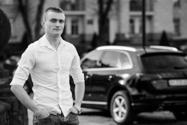 Beyaz tişörtlü genç adam lüks bir arabanın yanında güven içinde poz veriyor..