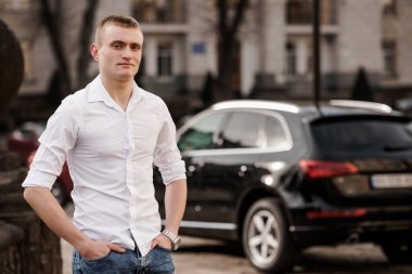 Beyaz Gömlekli Genç Adam Şehir Ayarları 'nda Lüks Siyah SUV' un yanında güven içinde poz veriyor..