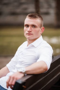 Beyaz tişörtlü genç bir adam açık havada bir bankta oturuyor ve düşünceli bir ifadeyle.