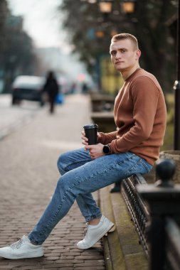 Parktaki bankta oturan genç bir adam, hayat dolu bir şehir ortamında kahve fincanıyla..