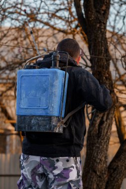 Güneş Batarken Bahçede Mavi Sırt Çantası Böcek İlacı Spreyi ile Koruyucu Giyinen Bir Kişinin Arka Görünümü.