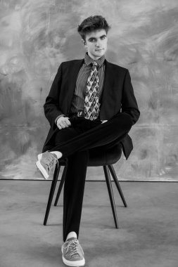 Takım elbiseli bir gencin siyah beyaz portresi. Ayakları sandalyenin üzerinde dokulu arka plana çarpık bir şekilde poz vermiş..