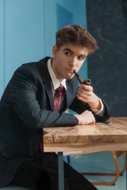Koyu takım elbiseli genç adam ahşap masaya yaslanırken dikkatlice pipo içiyor..