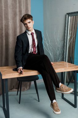 Resmi giyinmiş, siyah takım elbiseli, kırmızı kravatlı, rahat bir duruşu olan ve direk dik dik bakan genç bir adam..