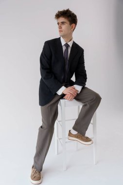 Resmî giyinmiş genç bir adam arka planda beyaz bir tabureye oturmuş, ellerini sıkarak dikkatlice uzaklara bakıyor..