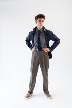 Mavili bir ceket, çizgili gömlek, gri pantolon ve beyaz arka planda spor ayakkabılardan oluşan kendine güvenen genç bir adam..