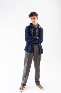 Kolları çapraz duran, mavi ceketli, çizgili kravatlı, gri pantolonlu ve beyaz arka planda sıradan ayakkabılı genç bir adam..