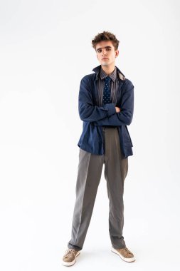 Kolları çaprazlanmış, katmanlı giysiler giymiş ceketi, gömleği, kravatı ve pantolonu beyaz bir arka planda duran genç bir adamın tüm vücut portresi..