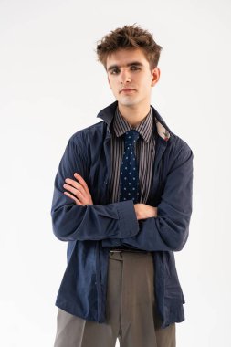 Kollarını kavuşturup stüdyoda poz veren genç adam çizgili gömlek, mavi ceket ve beyaz arka planda kravat giyiyor..